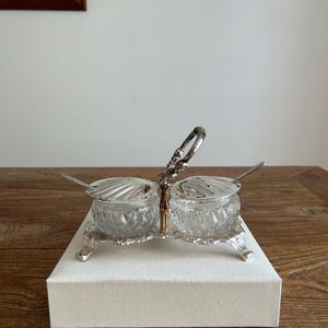 May include: A double condiment server featuring two clear, diamond-patterned glass bowls with silver-toned shell-shaped lids and matching spoons. The bowls are held in a silver-toned metal stand with a central decorative handle and four feet.
