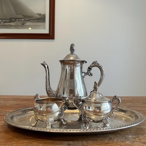 May include: A silver-plated tea service set consisting of a tall teapot, a lidded sugar bowl, and a creamer, all featuring ornate floral detailing on the handles and feet. The three pieces are arranged on a large, oval silver-plated serving tray.