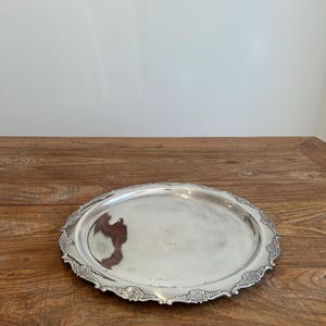 May include: A round silver-toned metal serving tray with a smooth, reflective centre surface and a decorative, raised border featuring intricate floral and leaf relief patterns along the scalloped edge of the circular platter.