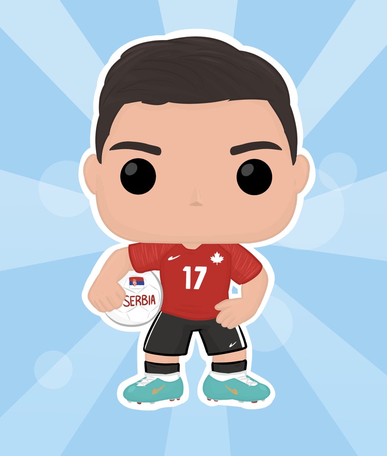 May include: A red and white soccer jersey with the number 17 on the front. The jersey has a Canadian flag patch on the right shoulder and a Nike logo on the left chest. The player is holding a soccer ball with the word "Serbia" and the Serbian flag on it.