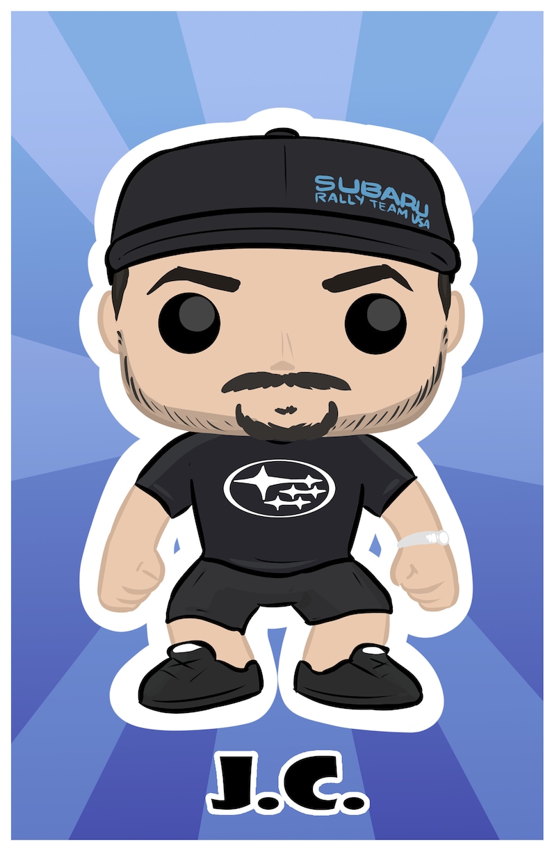 May include: A black and white pop culture figure wearing a black baseball cap with the text "SUBARU RALLY TEAM USA" on it. The figure is wearing a black t-shirt with a white Subaru logo. The figure is standing with its arms at its sides and has a black goatee. The figure is labeled "J.C."