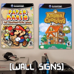May include: Two rectangular metal wall signs featuring Nintendo GameCube video game cover art. The left sign displays Paper Mario: The Thousand-Year Door, and the right sign displays Animal Crossing, both with ESRB E ratings and Nintendo logos.