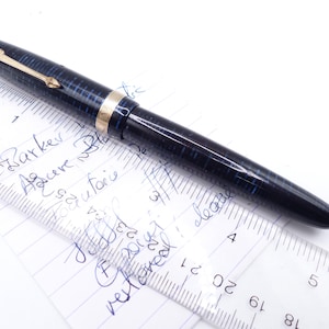May include: A vintage Parker fountain pen with a blue and black checkered pattern. The pen has gold-colored accents and a gold arrow-shaped clip. The pen is lying on a piece of paper with handwritten notes and a ruler.