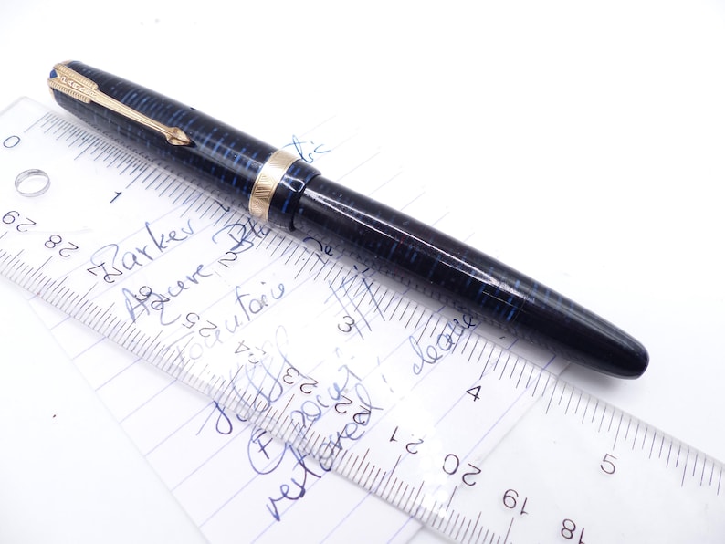 May include: A vintage Parker fountain pen with a blue and black checkered pattern. The pen has gold-colored accents and a gold arrow-shaped clip. The pen is lying on a piece of paper with handwritten notes and a ruler.