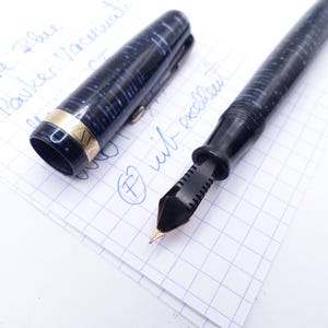 Op de afbeelding: Een zwarte en blauwe vulpen met een goudkleurige band en penpunt, liggend op een stuk ruitjespapier. De dop van de pen is eraf, waardoor de penpunt zichtbaar is. De woorden "Azare Blue Parker Vacumatic" zijn met blauwe inkt geschreven.