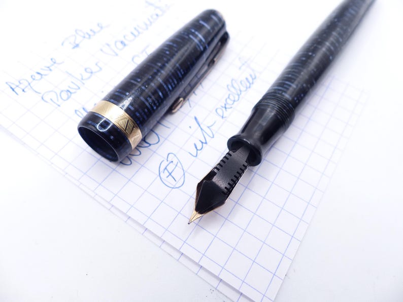 Op de afbeelding: Een zwarte en blauwe vulpen met een goudkleurige band en penpunt, liggend op een stuk ruitjespapier. De dop van de pen is eraf, waardoor de penpunt zichtbaar is. De woorden "Azare Blue Parker Vacumatic" zijn met blauwe inkt geschreven.
