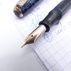 Op de afbeelding: Close-up van een vintage vulpen met een gouden penpunt. De pen heeft een zwarte houder met blauwe en gouden accenten op de dop. De penpunt ligt op ruitjespapier met blauwe inkt.