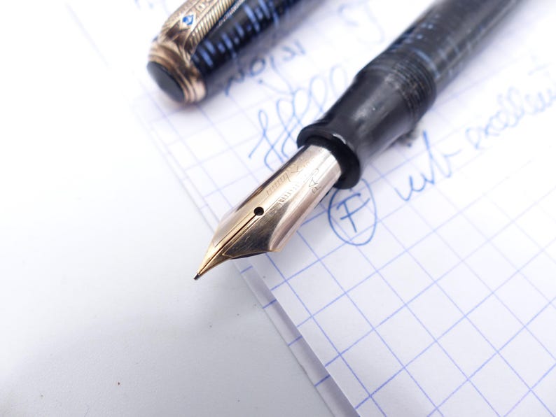 Op de afbeelding: Close-up van een vintage vulpen met een gouden penpunt. De pen heeft een zwarte houder met blauwe en gouden accenten op de dop. De penpunt ligt op ruitjespapier met blauwe inkt.