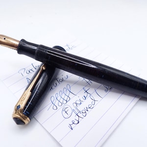 May include: A vintage fountain pen with a gold nib and a dark blue and black marbled barrel. The pen is partially disassembled, revealing the nib and the ink reservoir. The pen rests on a piece of paper with handwritten notes.
