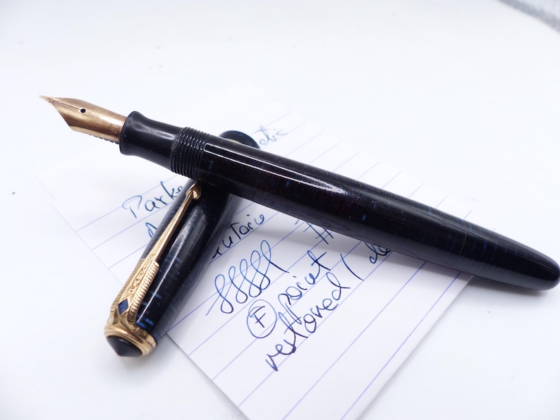 May include: A vintage fountain pen with a gold nib and a dark blue and black marbled barrel. The pen is partially disassembled, revealing the nib and the ink reservoir. The pen rests on a piece of paper with handwritten notes.