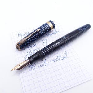 Op de afbeelding: Een vintage vulpen met een blauw en zwart gemarmerd ontwerp, gouden accenten en een gouden penpunt. De pen is open, waardoor de penpunt zichtbaar is, en ligt op een stuk ruitjespapier met handgeschreven notities. De dop van de pen is ook zichtbaar.