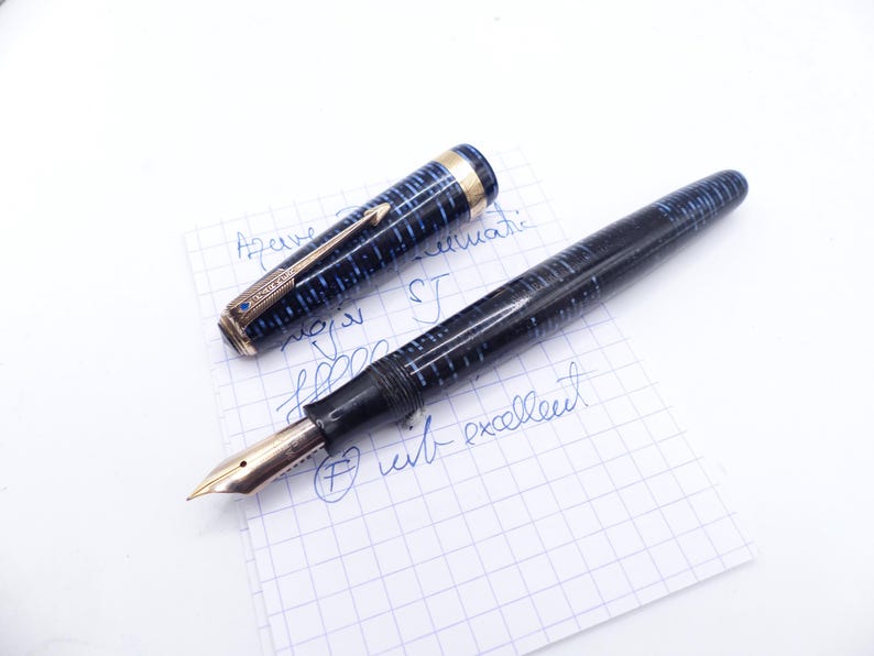 Op de afbeelding: Een vintage vulpen met een blauw en zwart gemarmerd ontwerp, gouden accenten en een gouden penpunt. De pen is open, waardoor de penpunt zichtbaar is, en ligt op een stuk ruitjespapier met handgeschreven notities. De dop van de pen is ook zichtbaar.