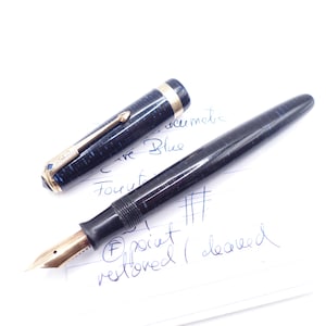May include: A black fountain pen with a gold nib and a detached cap. The cap is dark blue with gold accents. The pen is lying on a white surface with handwritten notes. The pen is labeled "Curve Blue".