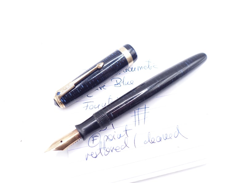 May include: A black fountain pen with a gold nib and a detached cap. The cap is dark blue with gold accents. The pen is lying on a white surface with handwritten notes. The pen is labeled "Curve Blue".