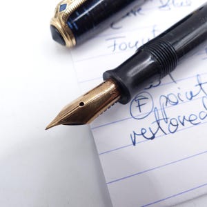 May include: Close-up of a vintage fountain pen with a gold nib and black barrel. The pen is partially disassembled, with the nib and section visible. The pen is resting on a lined paper with handwritten notes.