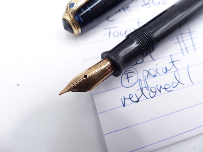 May include: Close-up of a vintage fountain pen with a gold nib and black barrel. The pen is partially disassembled, with the nib and section visible. The pen is resting on a lined paper with handwritten notes.