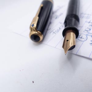 May include: A black fountain pen with a gold nib and accents, lying on a white surface next to its cap. The cap has gold trim and a black top. The nib is pointed towards the viewer.