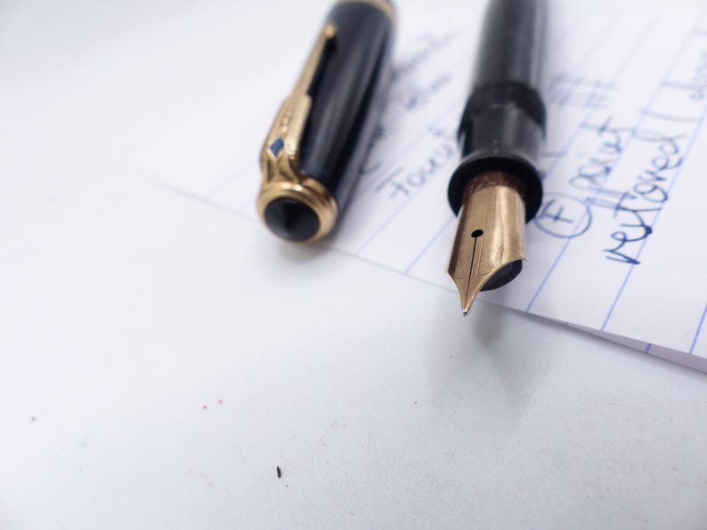 May include: A black fountain pen with a gold nib and accents, lying on a white surface next to its cap. The cap has gold trim and a black top. The nib is pointed towards the viewer.
