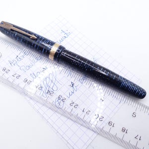 Op de afbeelding: Een vintage vulpen met een blauw en zwart gestreept patroon, gouden accenten en een goudkleurige clip. De pen ligt op een stuk ruitjespapier naast een liniaal met metingen in centimeters.