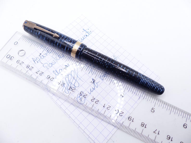 Op de afbeelding: Een vintage vulpen met een blauw en zwart gestreept patroon, gouden accenten en een goudkleurige clip. De pen ligt op een stuk ruitjespapier naast een liniaal met metingen in centimeters.