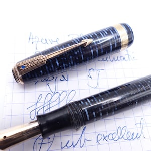 Op de afbeelding: Een vintage vulpen met een zwart en blauw patroon op de penhouder en goudkleurige accenten. De dop van de pen is eraf, waardoor de penpunt en het ingewikkelde ontwerp zichtbaar zijn. De pen ligt op geruit papier met blauwe inkt.