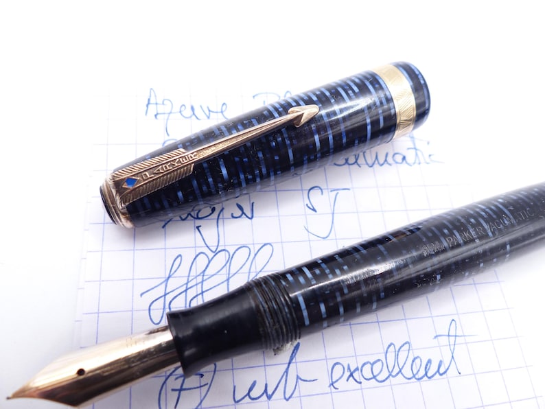 Op de afbeelding: Een vintage vulpen met een zwart en blauw patroon op de penhouder en goudkleurige accenten. De dop van de pen is eraf, waardoor de penpunt en het ingewikkelde ontwerp zichtbaar zijn. De pen ligt op geruit papier met blauwe inkt.