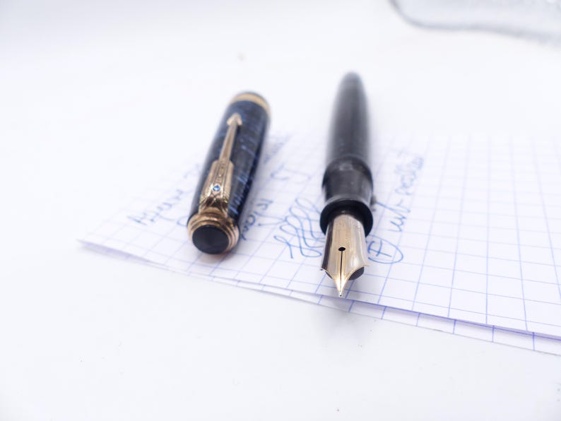 Op de afbeelding: Een zwarte vulpen met een gouden penpunt en een losse dop. De dop is donkerblauw met gouden details. De pen ligt op een stuk ruitjespapier met blauwe inktmarkeringen.