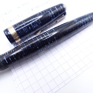 Op de afbeelding: Een vintage vulpen met een zwart-blauw gespikkeld ontwerp. De dop is eraf, met een gouden band en een zilveren clip. De pen ligt op een wit oppervlak met een rasterpatroon, wat wijst op een schrijf- of tekencontext.