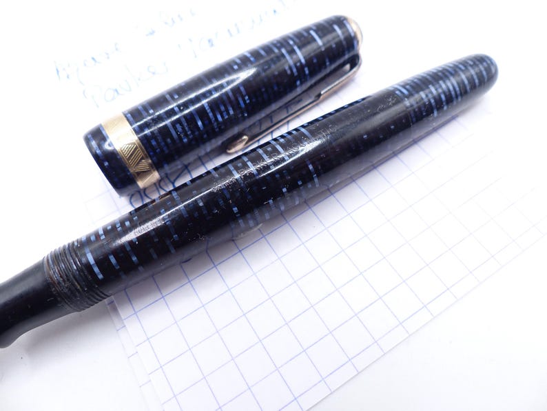 Op de afbeelding: Een vintage vulpen met een zwart-blauw gespikkeld ontwerp. De dop is eraf, met een gouden band en een zilveren clip. De pen ligt op een wit oppervlak met een rasterpatroon, wat wijst op een schrijf- of tekencontext.