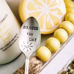 Puede incluir: Una cuchara de plata con las palabras "Squeeze the Day" grabadas. La cuchara est&aacute; rodeada de cuentas de madera amarillas y un adorno de cer&aacute;mica con forma de lim&oacute;n.