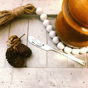 May include: A silver butter knife with the text "HEY HONEY" and a bee graphic. The knife is on a white wooden surface, next to pinecones, a brown jar, and white beads.