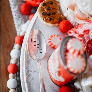 May include: A silver spoon with the words "HO HO HO" engraved on it. The spoon is on a white wooden tray with red and white candy, a cookie, and a red and white can.