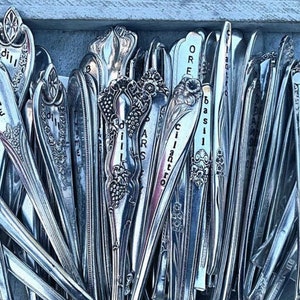 May include: A collection of vintage silverware spoons with engraved names of herbs, including oregano, thyme, basil, cilantro, and dill. The spoons are arranged in a vertical row, with the names facing the viewer.