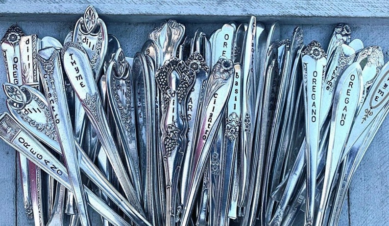 May include: A collection of vintage silverware spoons with engraved names of herbs, including oregano, thyme, basil, cilantro, and dill. The spoons are arranged in a vertical row, with the names facing the viewer.