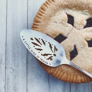 May include: A silver pie server with a decorative design and the text "A Little Slice of Heaven" is resting on top of a homemade pie with a lattice crust and a dark purple filling.