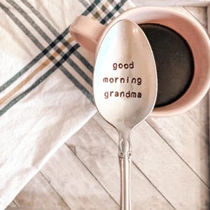 May include: A silver spoon with the words "good morning grandma" stamped on the bowl. The spoon is next to a pink mug with dark liquid inside, resting on a white and plaid cloth.