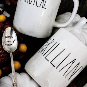 May include: Two white ceramic mugs with black lettering. One mug says "MAGICAL" and the other says "BRILLIANT". A silver spoon with the words "ACCIO HOT COCOA" is in the foreground.
