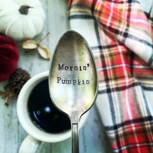 May include: A silver spoon with the words "Morning Pumpkin" engraved on the front. The spoon is sitting on a white surface with a plaid blanket and a cup of coffee in the background.