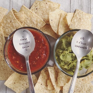 May include: Two silver spoons with engraved sayings. One spoon says "Nacho Ordinary Salsa" and the other says "It's 5 O'Clock Somewhere". The spoons are in bowls of salsa and guacamole, surrounded by tortilla chips.