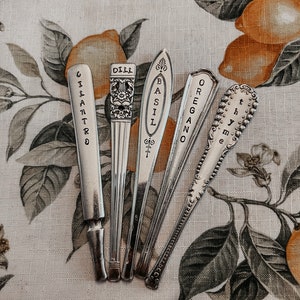 May include: Five silver spoons with engraved labels for different herbs: Cilantro, Dill, Basil, Oregano, and Thyme. The spoons are arranged on a floral patterned fabric with a lemon motif.