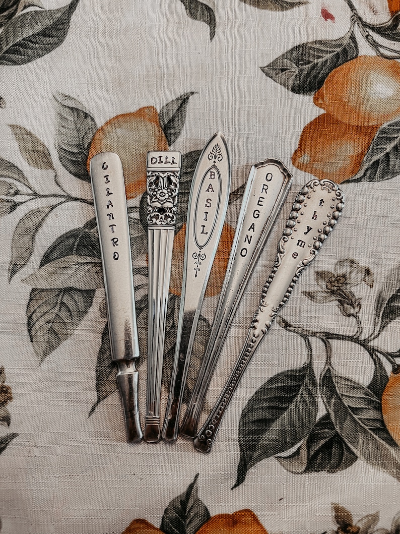 May include: Five silver spoons with engraved labels for different herbs: Cilantro, Dill, Basil, Oregano, and Thyme. The spoons are arranged on a floral patterned fabric with a lemon motif.