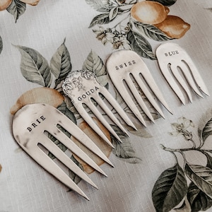 May include: Four silver cheese forks with the names "BRIE", "GOUDA", "SWISS", and "BLUE" stamped on them. The forks are arranged on a floral patterned tablecloth with lemons and leaves.