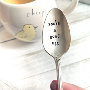 May include: A silver spoon with the words "you're a good egg" engraved on it. The spoon is being held by a hand with pink nail polish.