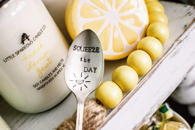 Puede incluir: Una cuchara de plata con las palabras "Squeeze the Day" grabadas. La cuchara est&aacute; rodeada de cuentas de madera amarillas y un adorno de cer&aacute;mica con forma de lim&oacute;n.
