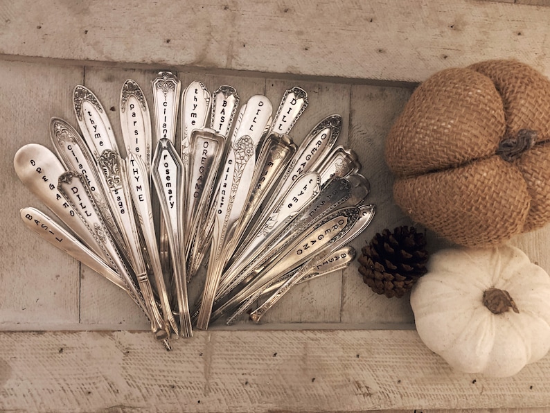 May include: A collection of vintage silverware spoons with various herbs and spices names engraved on them. The spoons are arranged in a fan shape on a wooden surface. There is a burlap pumpkin and a white pumpkin in the background.