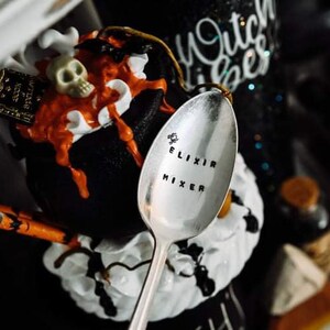 May include: A silver spoon with the words "ELIXIR MIXER" stamped on it. The spoon is in front of a black cauldron with orange liquid, a skull, and a small book. The background includes a bottle and the words "Witch Vibes".