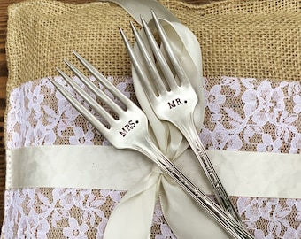 May include: Two silver forks with the words "MRS." and "MR." engraved on the handles. The forks are placed on a burlap pillow with white lace trim and a white satin ribbon.