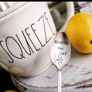 Puede incluir: Una jarra de cer&aacute;mica blanca con la palabra "SQUEEZE" escrita en ella, una cuchara de plata con las palabras "SQUEEZE the DAY" grabadas en ella y un lim&oacute;n amarillo.