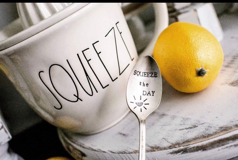 Puede incluir: Una jarra de cer&aacute;mica blanca con la palabra "SQUEEZE" escrita en ella, una cuchara de plata con las palabras "SQUEEZE the DAY" grabadas en ella y un lim&oacute;n amarillo.