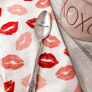 May include: A silver teaspoon with the inscription "p.s. i love you" on a white background with pink and red lips printed on it.